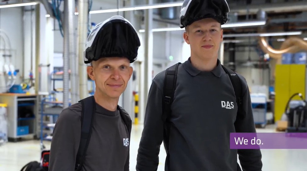 Two individuals wearing welding helmets and carrying backpacks stand inside a well-lit industrial manufacturing facility, featuring various equipment and machinery in the background.
