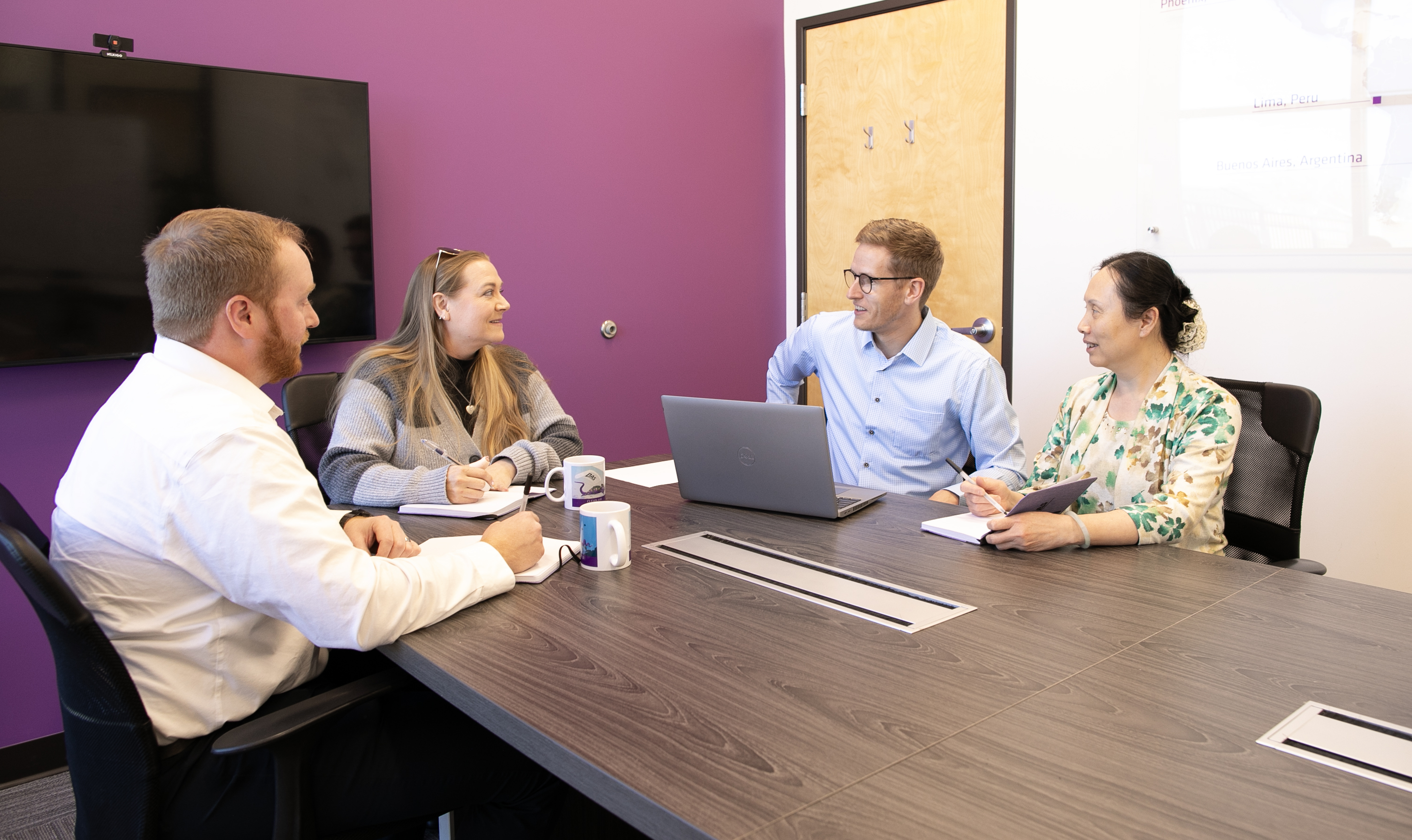 A group of individuals sitting around a conference table with cups, laptop und notebooks on the table, engaging in a conversation