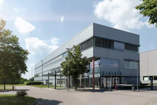 AMTC Dresden A modern, rectangular office building with a sleek, metallic facade and large windows, surrounded by a spacious, tree-lined street under a partly cloudy sky.