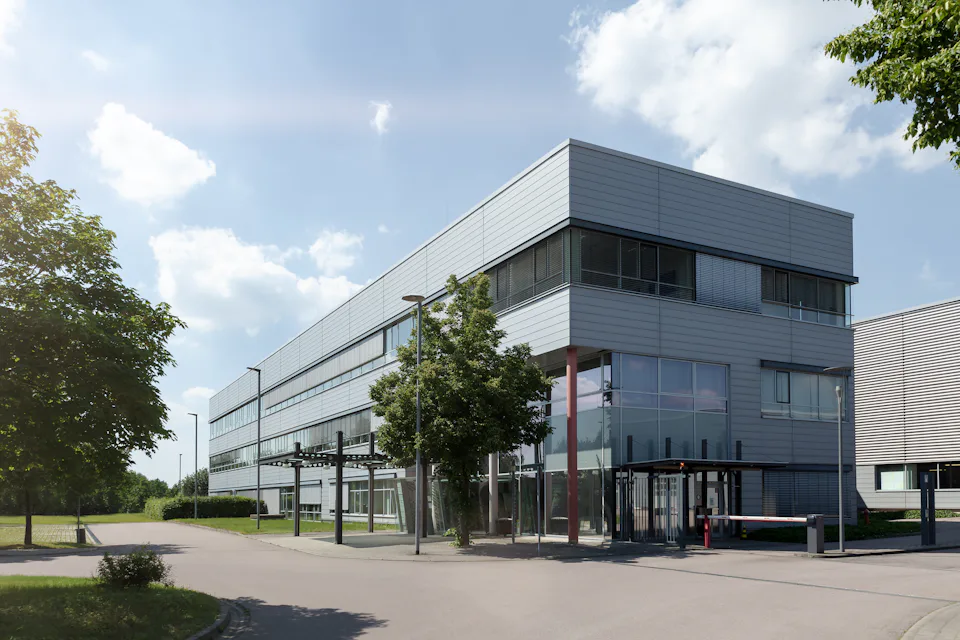 AMTC Dresden A modern, rectangular office building with a sleek, metallic facade and large windows, surrounded by a spacious, tree-lined street under a partly cloudy sky.