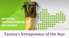 Award Saxony's Entrepreneur of the Year 이 이미지에는 양팔을 머리 위로 들어 올린 인물의 청동상이 담겨 있으며, 녹색 그라데이션과 픽셀 처리된 작센 주 지도가 배경으로 깔려 있고, “Sachsens Unternehmer:in des Jahres”라는 문구가 함께 적혀 있어 ‘작센 주 올해의 기업가’ 상을 상징하고 있습니다.