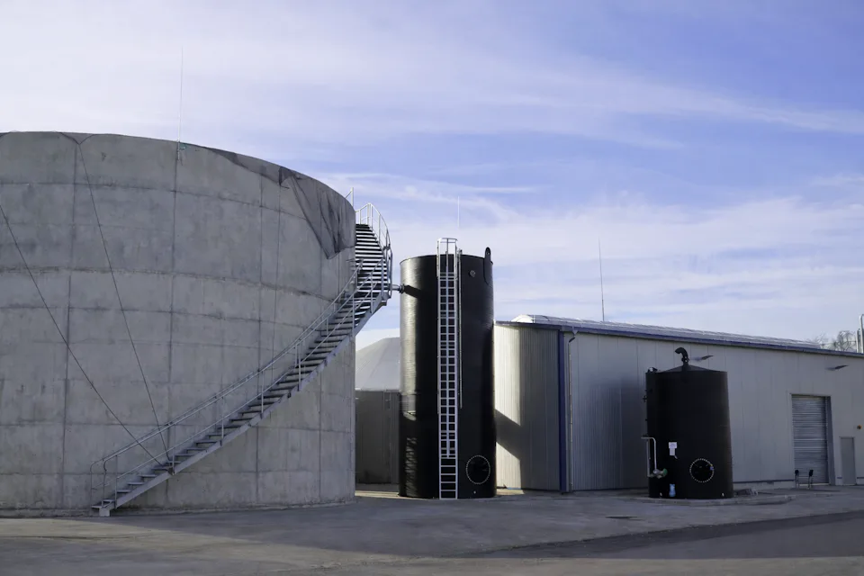 A large, cylindrical concrete storage tank with a metal staircase is situated next to two smaller, vertical black tanks and an industrial building under a clear blue sky, showcasing industrial architecture and infrastructure.
