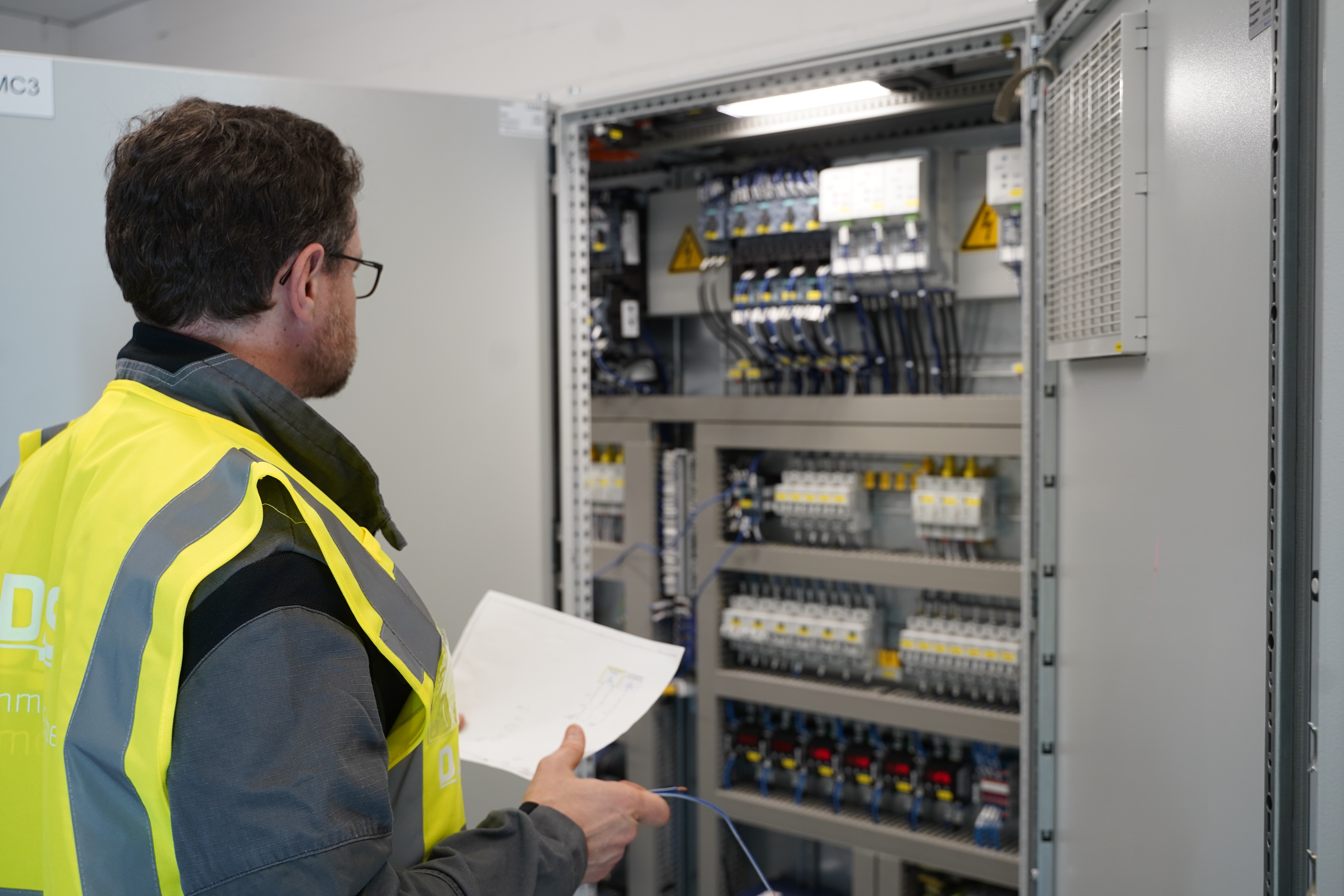 A technician wearing a yellow safety vest examines and takes notes on an open electrical panel filled with various switches and wiring in a well-lit industrial setting.