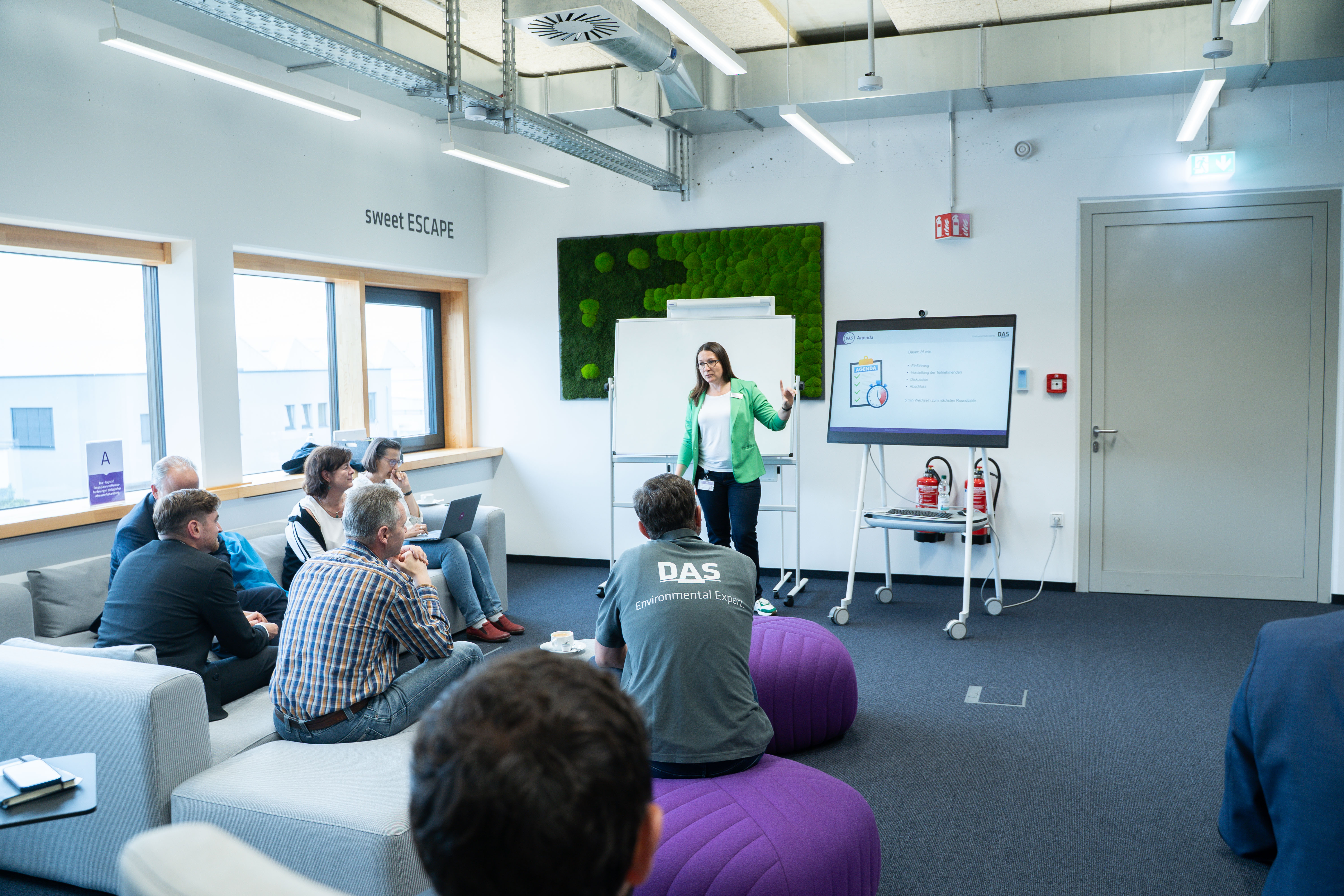 A group of professionals sits in an office meeting room with large windows, as a woman stands near a digital screen presenting data; modern office furniture and a green wall accent are featured.