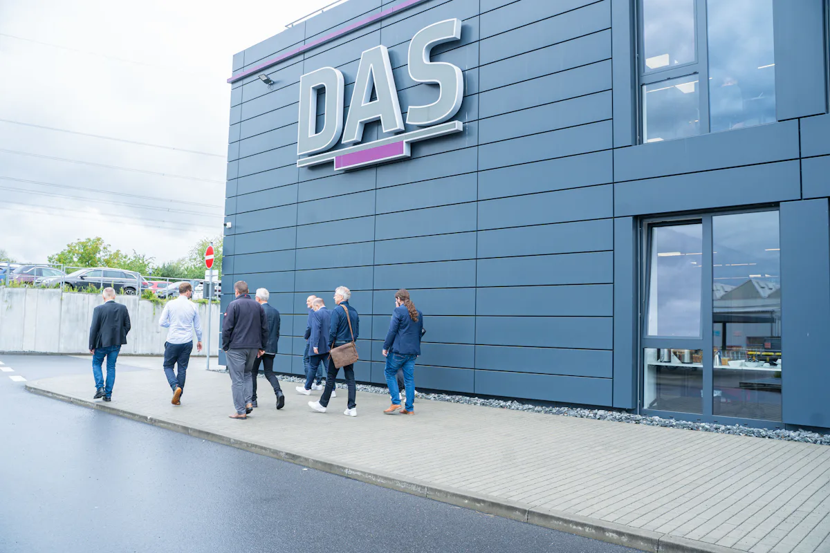 Impression Industrial Water Day 2025 A group of six people walk along a wet pavement outside a modern, dark gray building with large "DAS" signage, under an overcast sky.