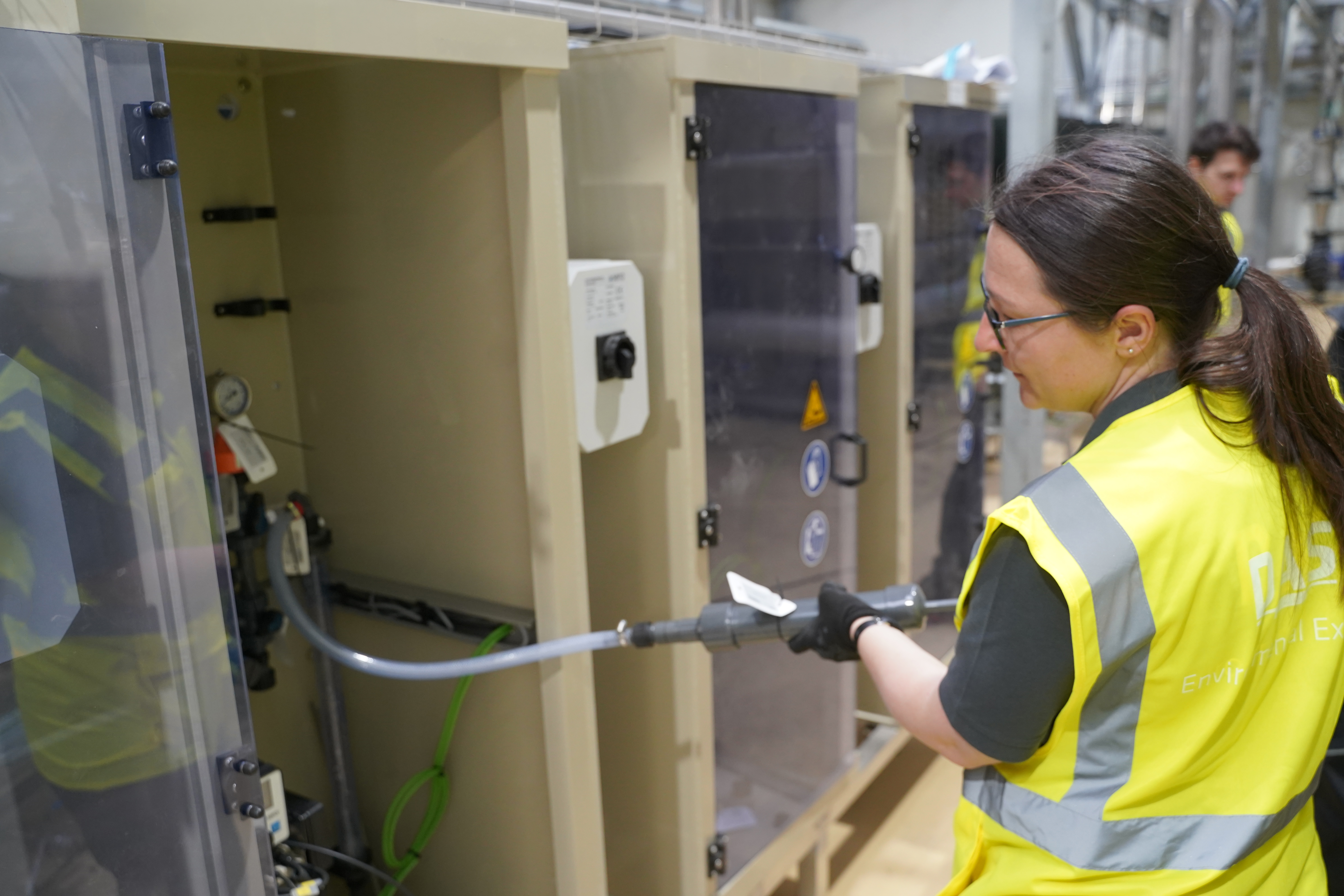 A person wearing a yellow safety vest is making adjustments to a large industrial control cabinet, surrounded by numerous cables and displays.