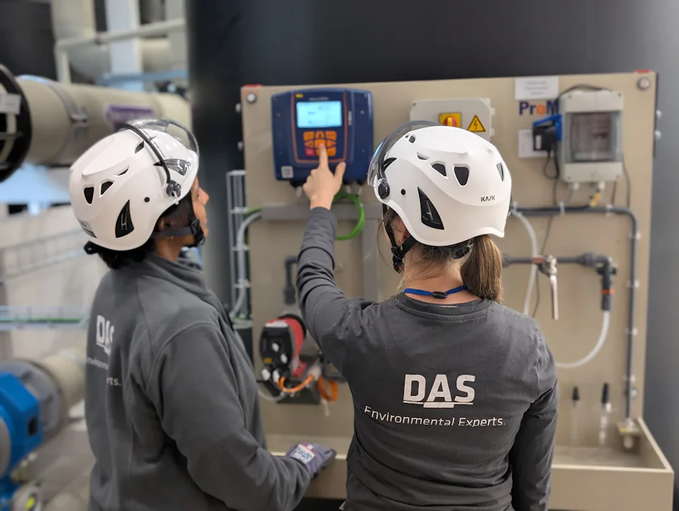 Two individuals wearing helmets and DAS-branded gear operate a sophisticated control panel in an industrial setting, highlighting environmental technology solutions.
