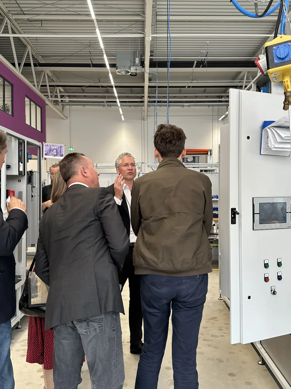 Press Visit June 2026_Tour of the Production Facility A group of people is standing in a modern, well-lit factory space, looking at a complex machine; the image depicts manufacturing processes.