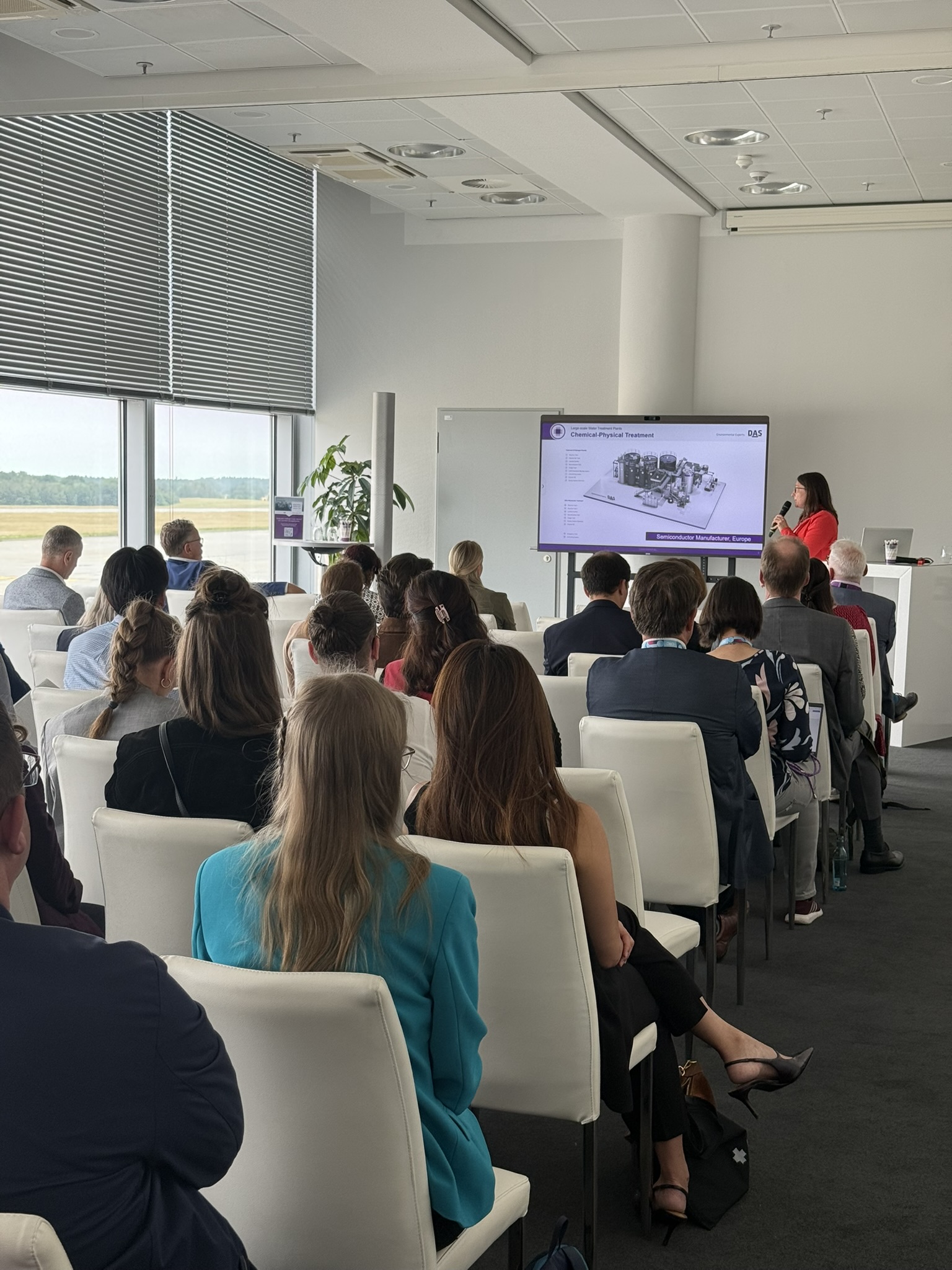 A group of people seated in a modern conference room attentively listens to a presentation on a large screen displaying architectural blueprints.