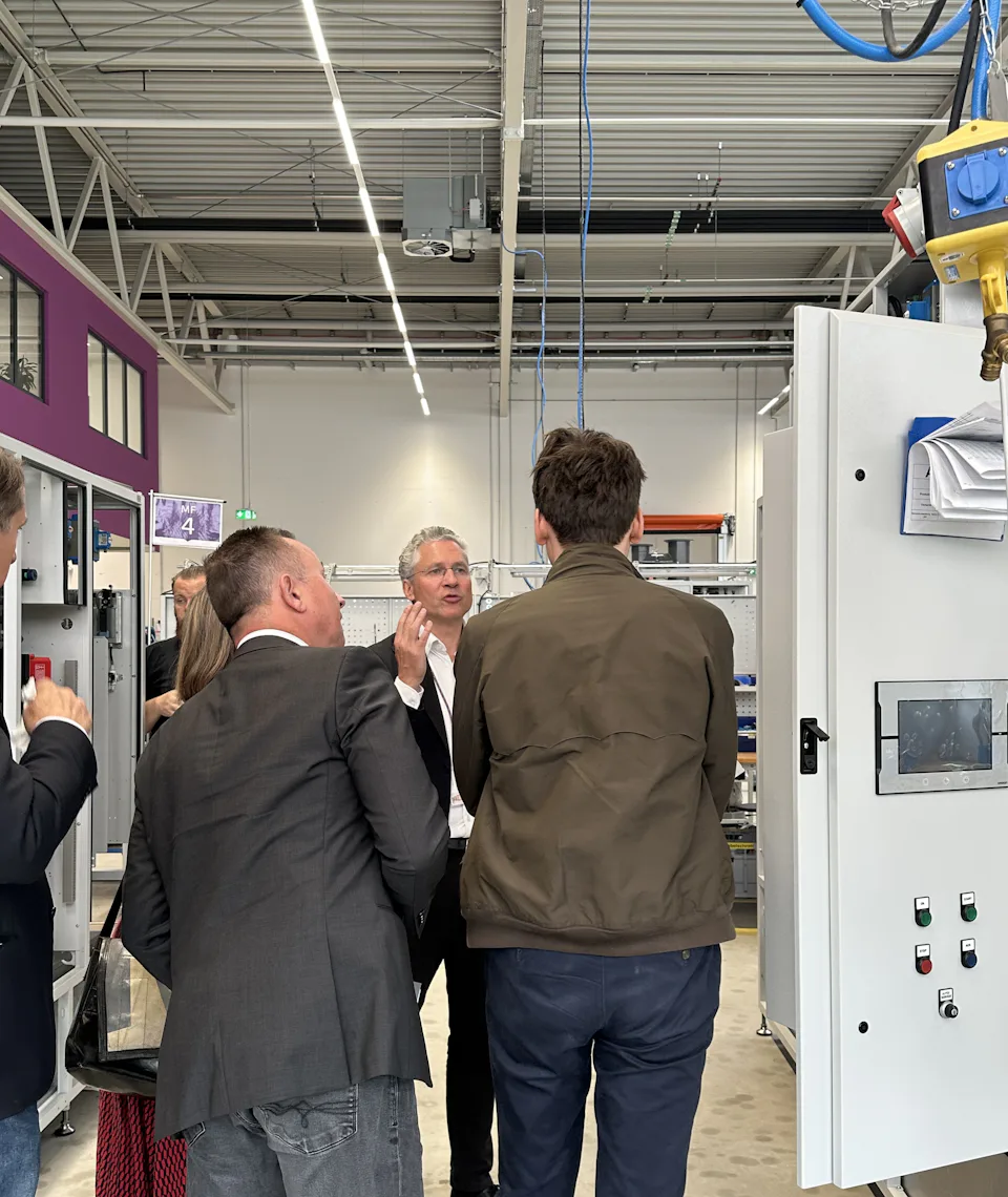 A group of people is standing in a modern, well-lit factory space, looking at a complex machine; the image depicts manufacturing processes.