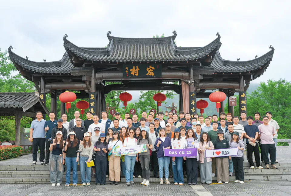 25th Anniversary DAS China A large group of people stands in front of a traditional Chinese wooden archway adorned with red lanterns, surrounded by lush greenery, with many holding colorful banners celebrating DAS China 25.