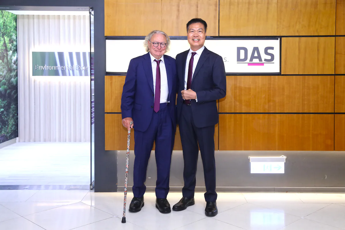 Dr. Horst Reichardt and Alex 合影 兩位穿著正裝的商務人士站在辦公室大樓內,其中一人手握雕花手杖,背景是一個帶有「DAS」和「I Environnement」標誌的木牆。