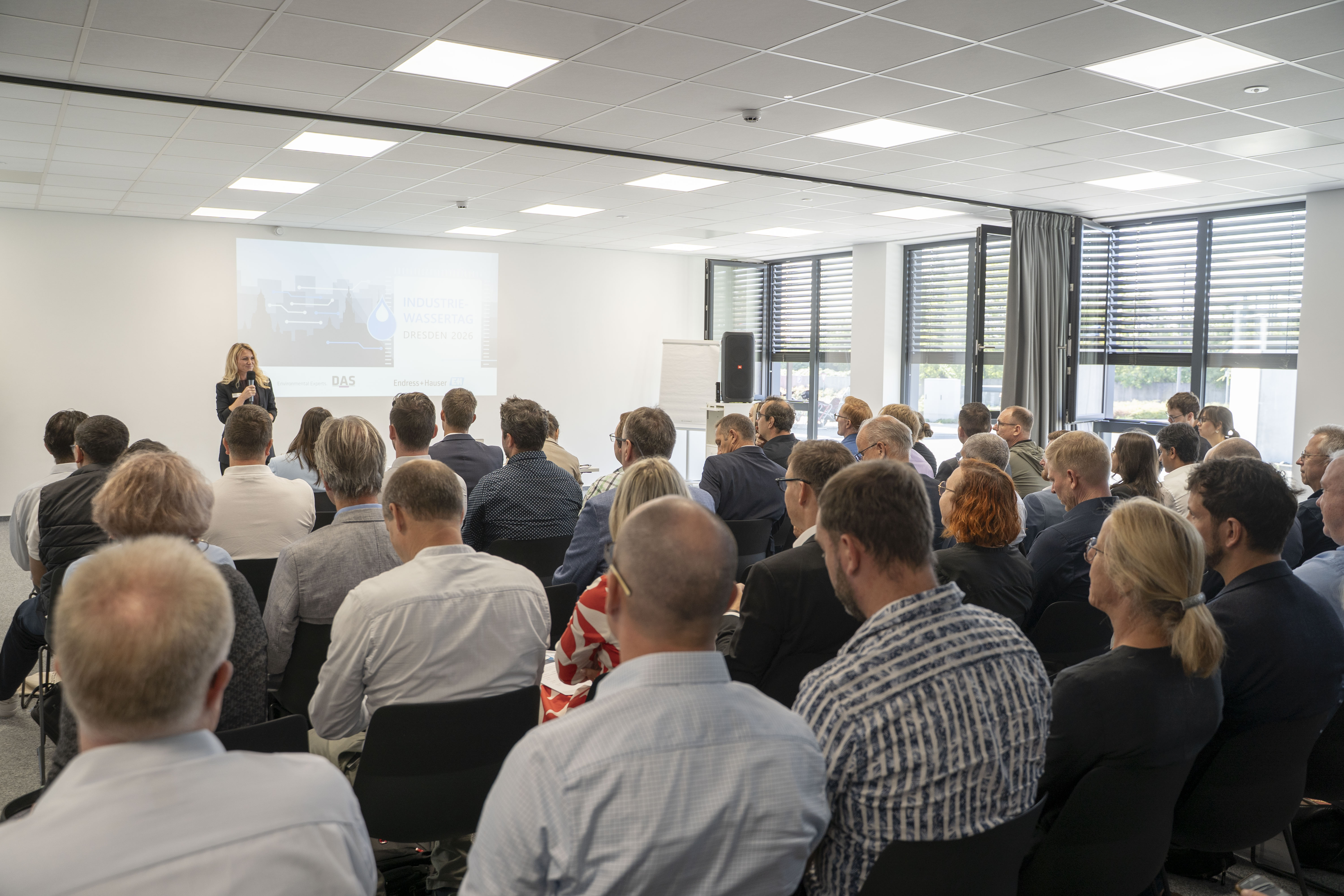 Eine große Gruppe von Menschen sitzt in einem Konferenzraum und hört einer Vortragenden vor einer Präsentation auf einer Leinwand zu, während natürliches Licht durch große Fenster einfällt.