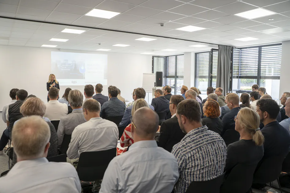Industriewassertag Dresden 2026 Eine große Gruppe von Menschen sitzt in einem Konferenzraum und hört einer Vortragenden vor einer Präsentation auf einer Leinwand zu, während natürliches Licht durch große Fenster einfällt.