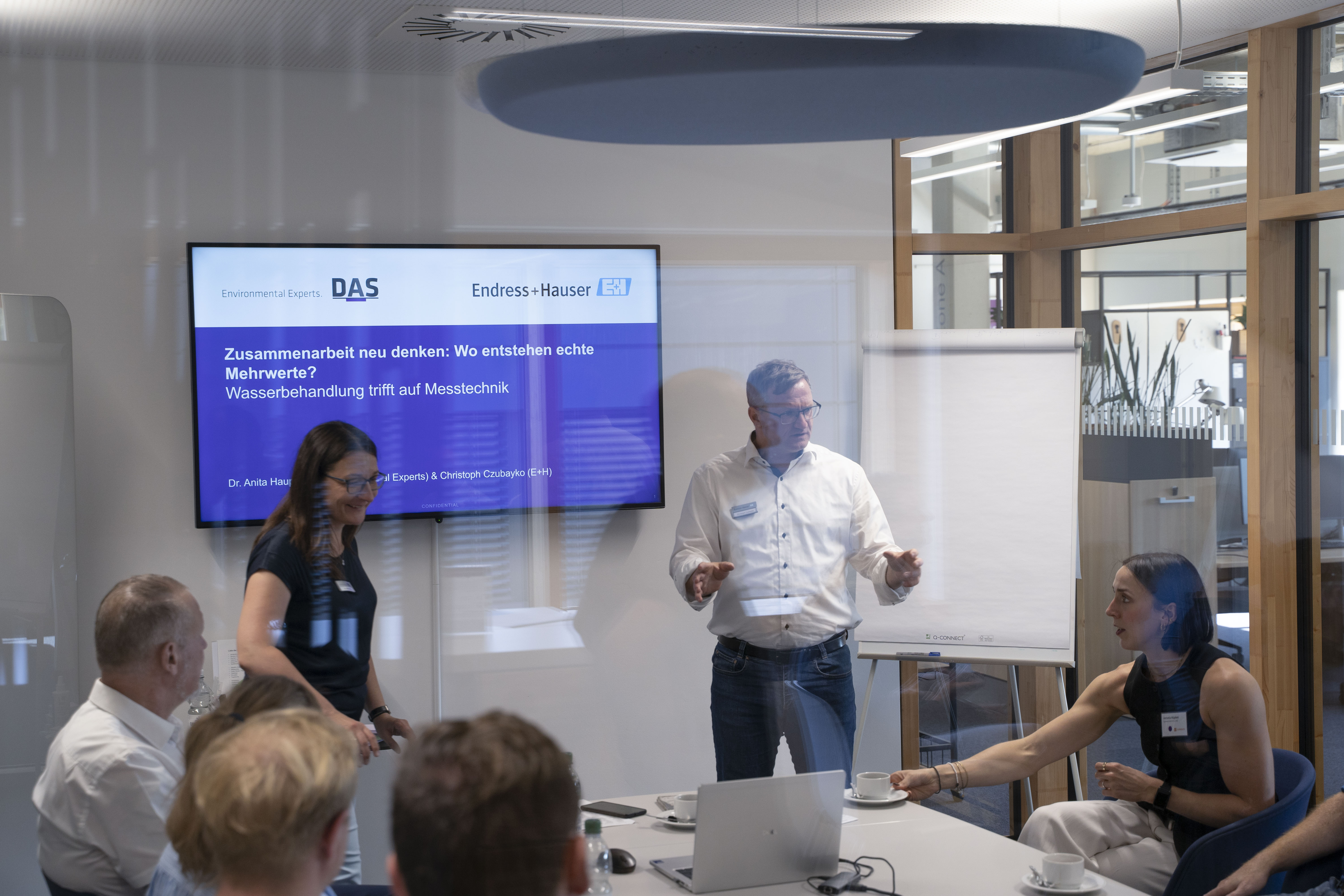 Eine Gruppe von Menschen sitzt in einem modernen Konferenzraum mit Glaswänden; auf einem großen Bildschirm sind Präsentationsfolien zur Wasserbehandlung und Messtechnik zu sehen, während ein Mann am Whiteboard steht und diskutiert.