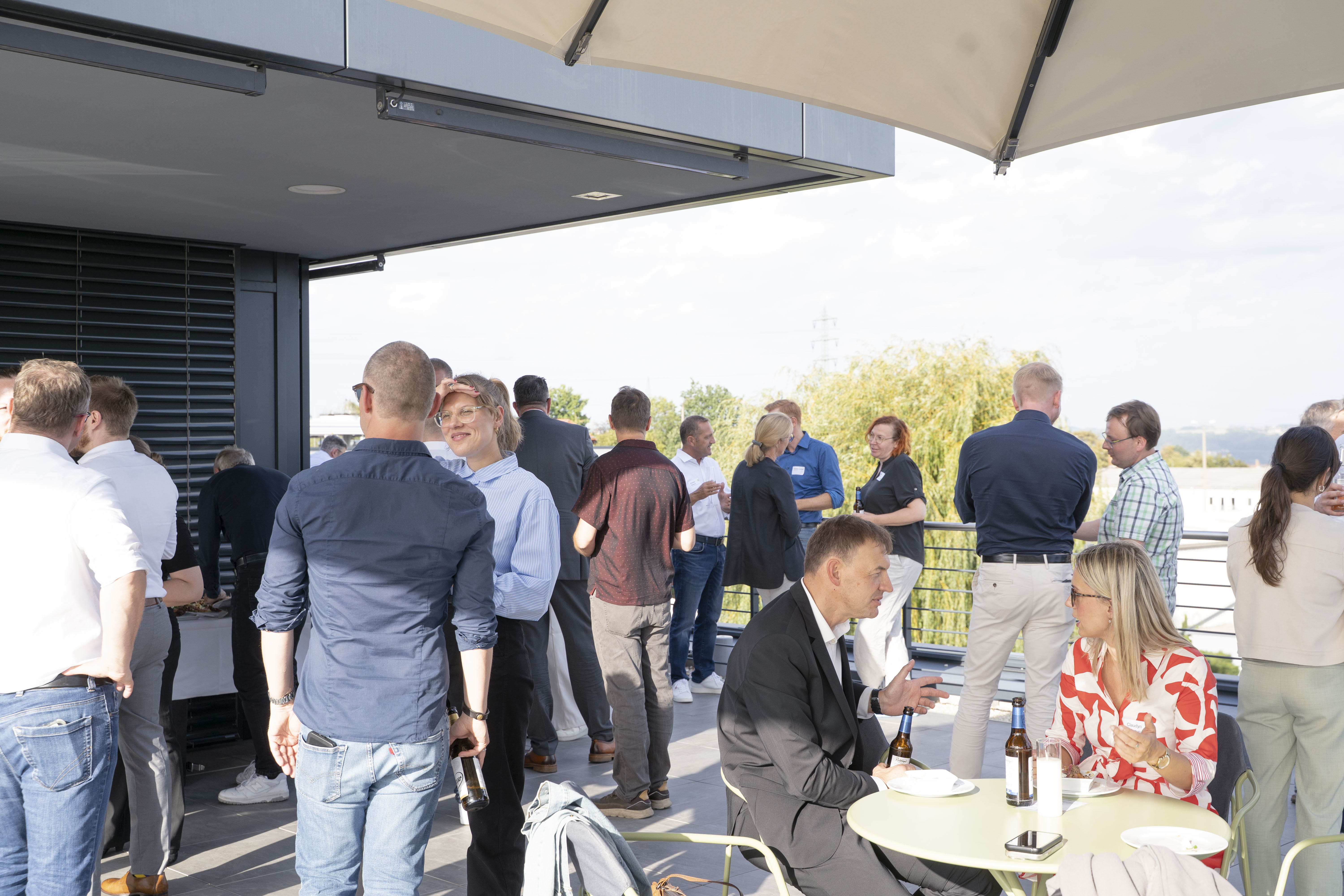Eine Gruppe von Menschen unterhält sich bei einer entspannten Veranstaltung im Freien auf einer Terrasse, wo einige Teilnehmer stehen und Getränke an kleinen Tischen genießen.