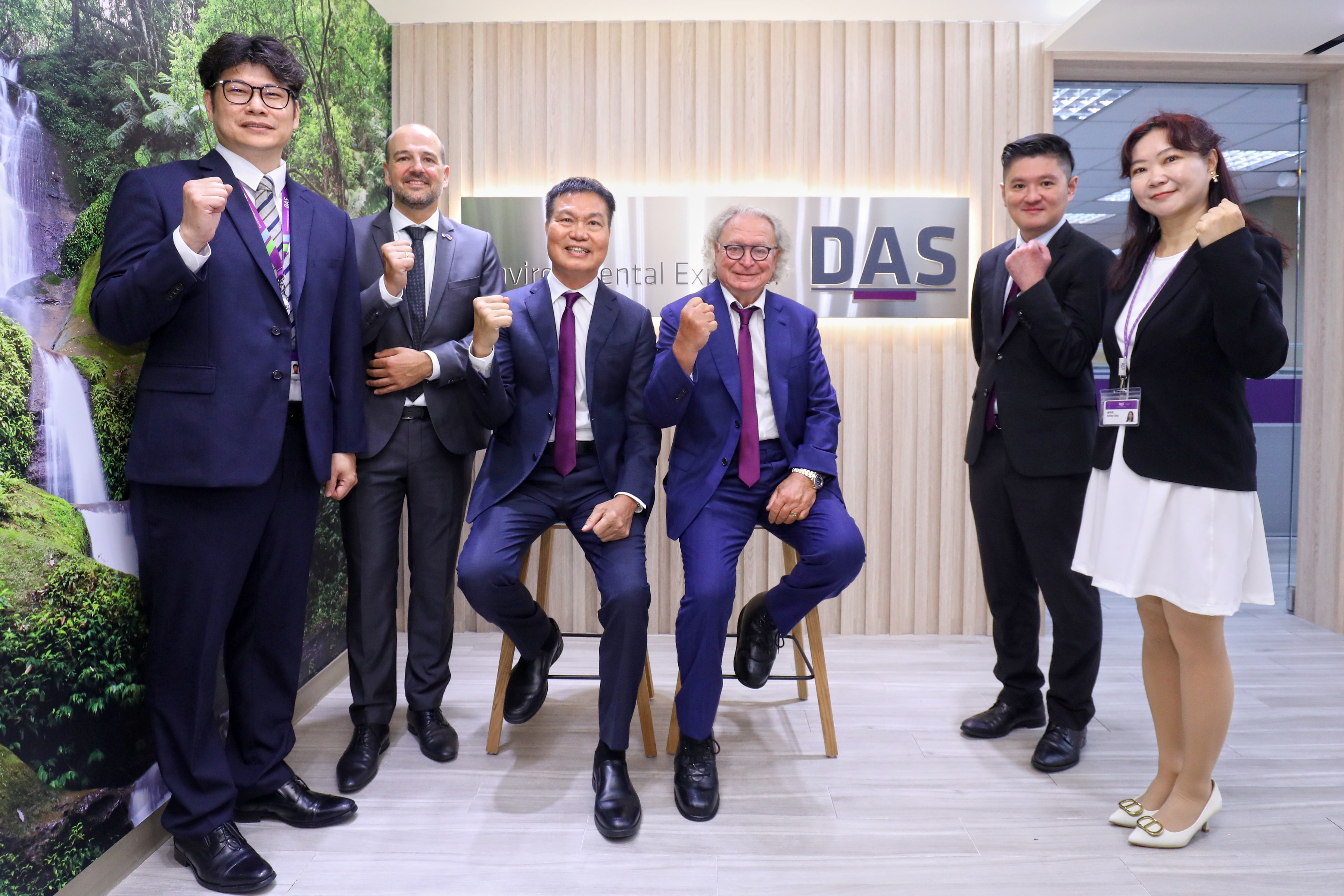 圖片顯示六位穿著正式商務裝的人在專業的辦公環境中，背景牆上有公司標誌，其中一人坐著，其他人站在旁邊；大家擺出握拳姿勢，展現團結與自信。