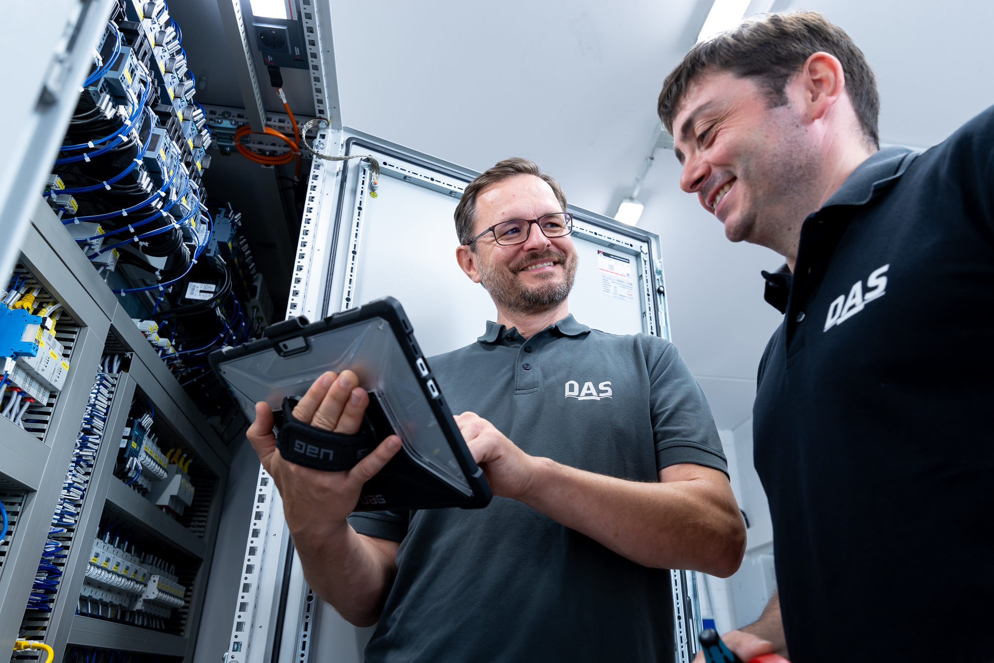 Two men in a technical room are examining equipment; one holds a tablet and they both wear matching branded polo shirts, surrounded by complex wiring and electrical panels.