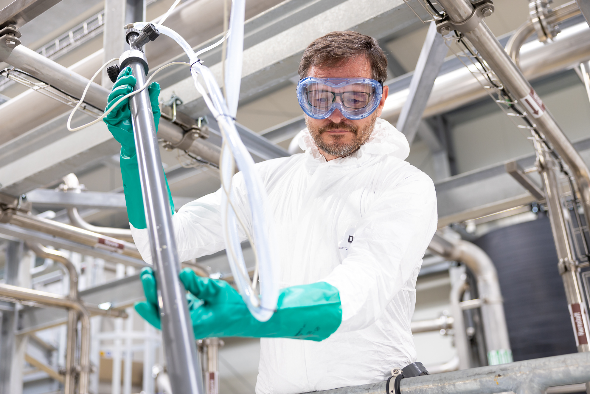 An engineer in a white protective suit and blue goggles works carefully with industrial piping, surrounded by stainless steel equipment, in a modern industry setting, highlighting industrial safety protocols.