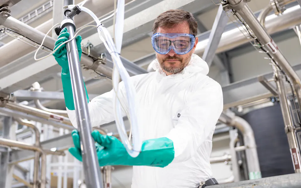 DAS Engineer Probing Wastewater An engineer in a white protective suit and blue goggles works carefully with industrial piping, surrounded by stainless steel equipment, in a modern industry setting, highlighting industrial safety protocols.