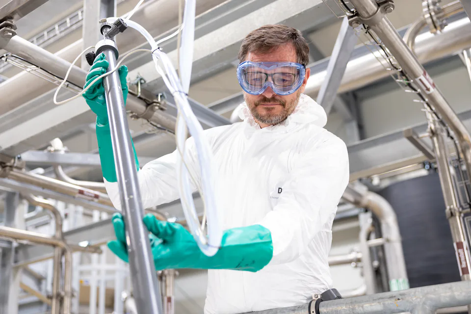 An engineer in a white protective suit and blue goggles works carefully with industrial piping, surrounded by stainless steel equipment, in a modern industry setting, highlighting industrial safety protocols.