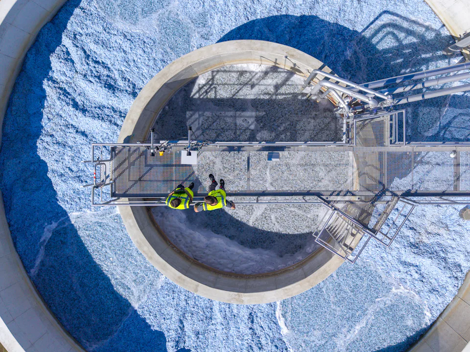 Aerial view of two engineers in high-visibility clothing inspecting a circular wastewater treatment facility with foamy water, highlighting industrial water management and environmental engineering.