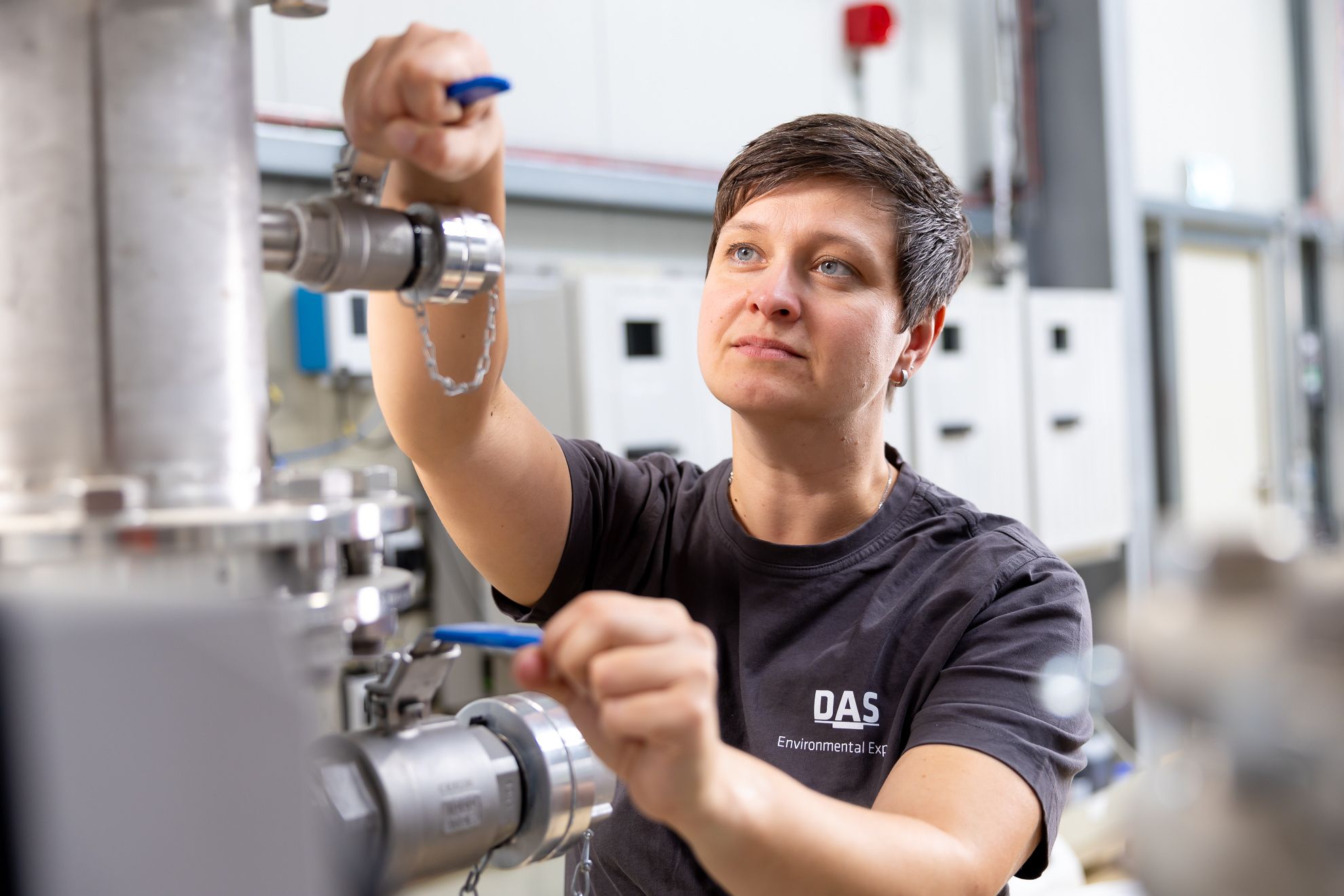 A technician in a workshop adjusts metal valves on industrial pipes, wearing a DAS Environmental shirt while concentrating on the task amidst technical equipment.
