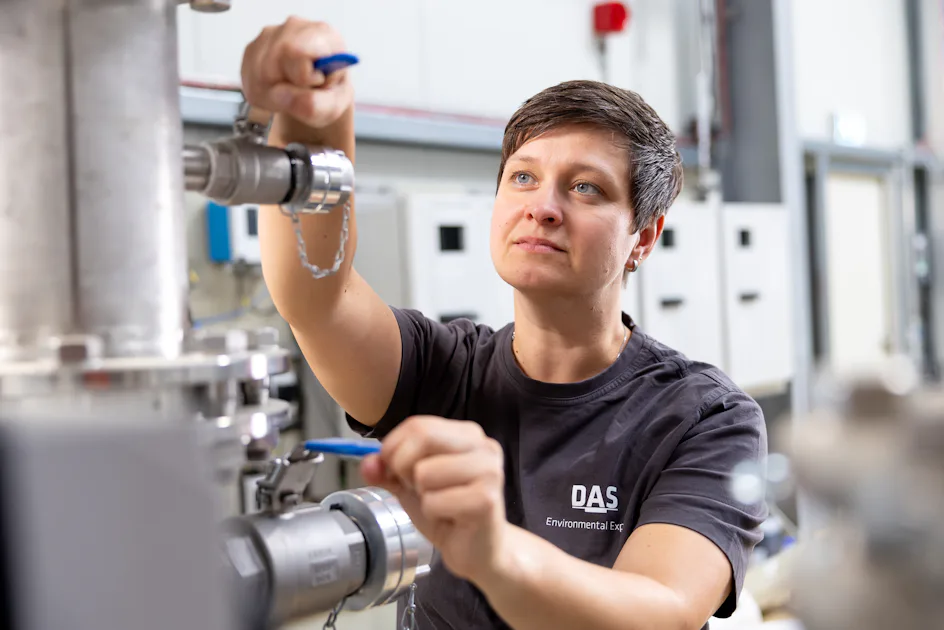 DAS Engineer adjusting valves A technician in a workshop adjusts metal valves on industrial pipes, wearing a DAS Environmental shirt while concentrating on the task amidst technical equipment.