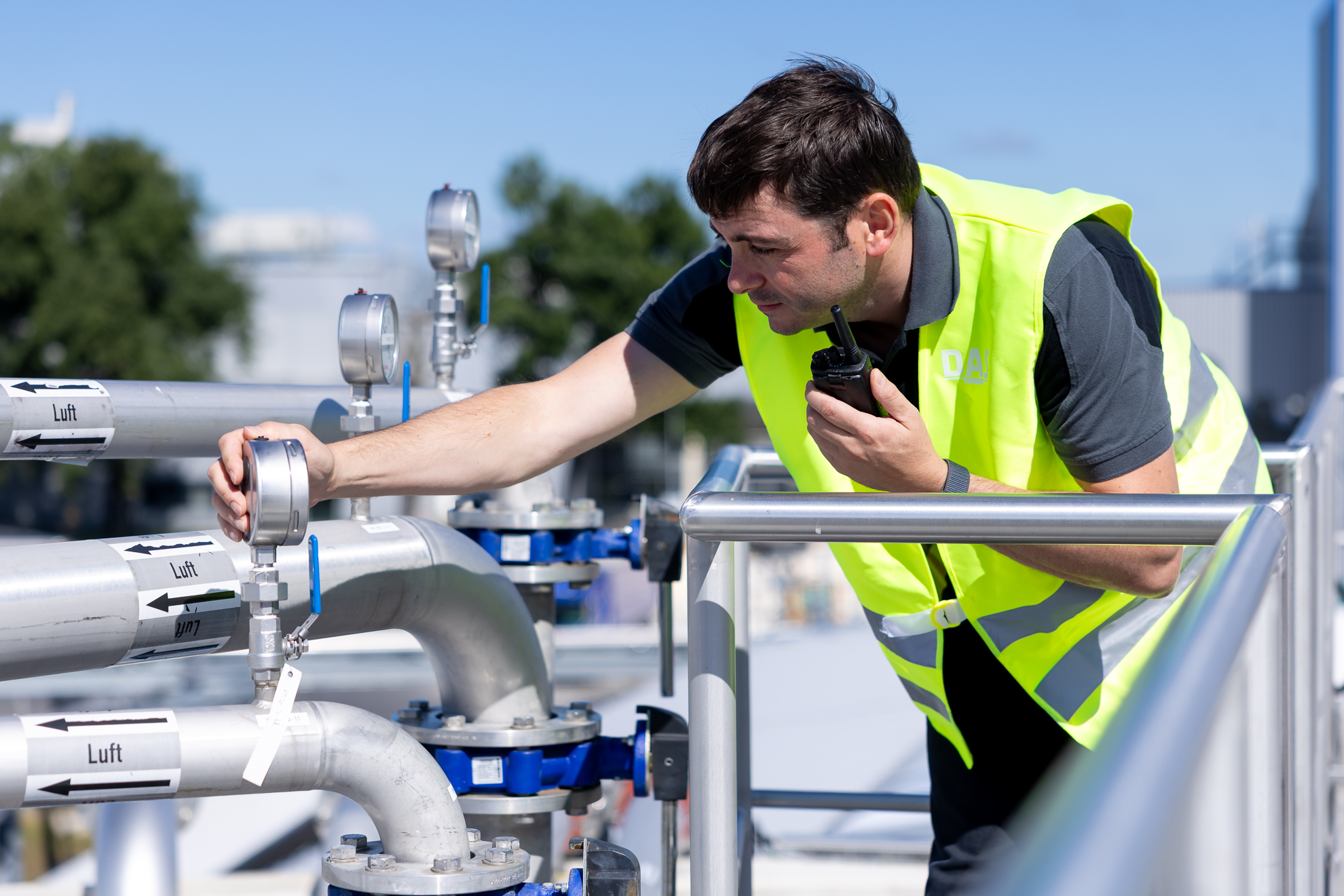 A worker wearing a reflective vest inspects pressure gauges on industrial pipes under a clear blue sky, ensuring operational safety and functionality.
