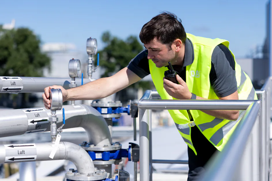 Emsland Aller Aqua Male DAS Engineer A worker wearing a reflective vest inspects pressure gauges on industrial pipes under a clear blue sky, ensuring operational safety and functionality.