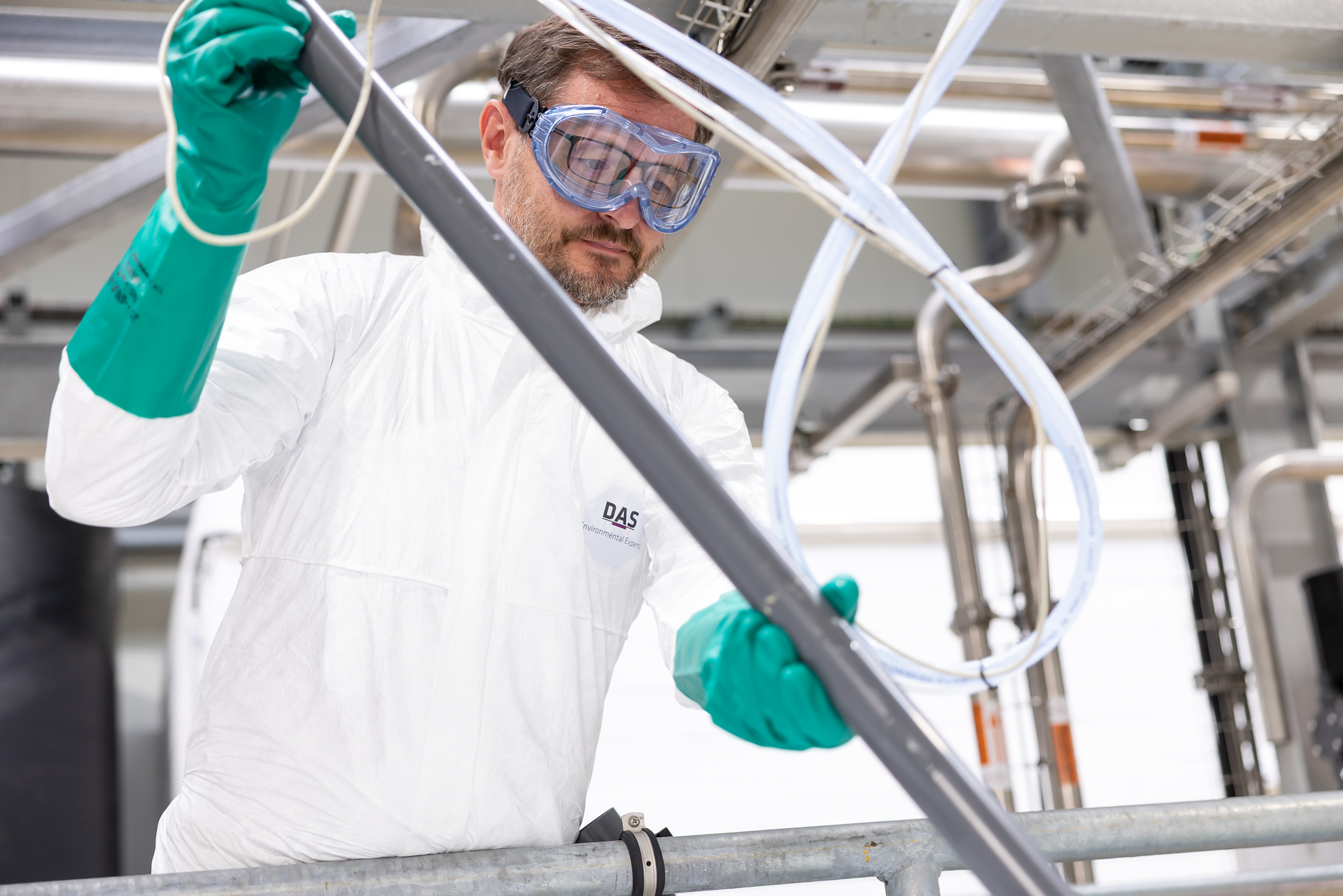 An engineer in a white protective suit and blue goggles works carefully with industrial piping, surrounded by stainless steel equipment, in a modern industry setting, highlighting industrial safety protocols.