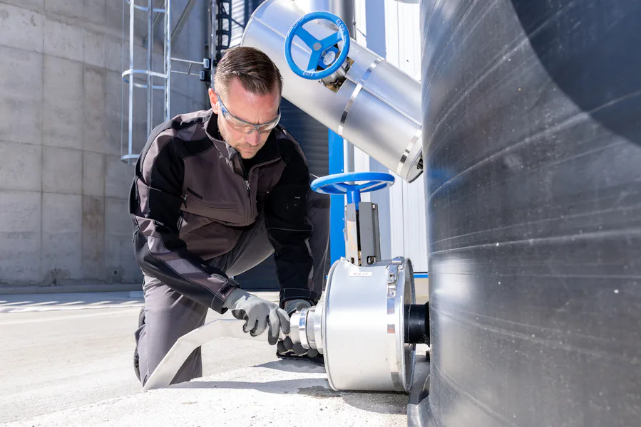 Emsland Aller Aqua DAS Worker A worker in safety gear kneels on concrete while fastening a large metal valve to a black industrial tank, showcasing industrial safety procedures in an outdoor setting.