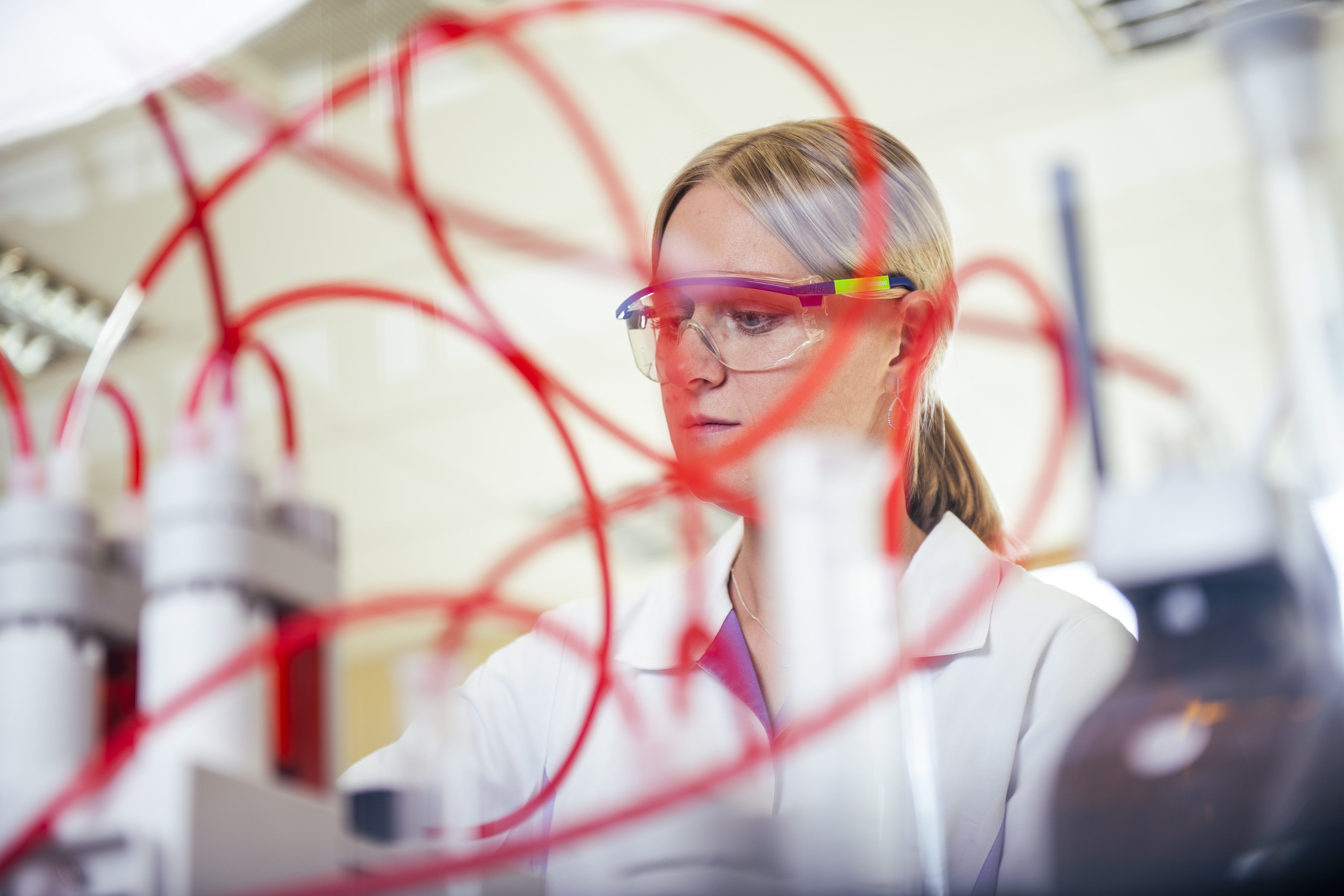 Eine Wissenschaftlerin in einem Labor, die eine Schutzbrille trägt, arbeitet mit komplizierten roten Schläuchen und hochmodernen wissenschaftlichen Geräten und konzentriert sich ganz auf die Durchführung eines Experiments.