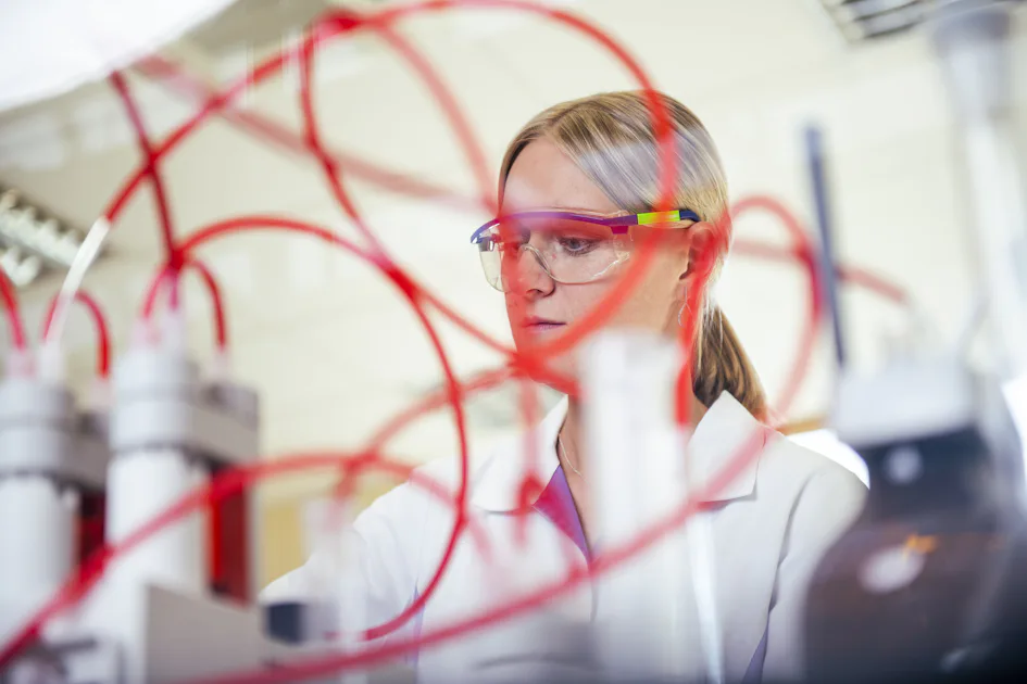 Labormitarbeitende an Analyseanlage Eine Wissenschaftlerin in einem Labor, die eine Schutzbrille trägt, arbeitet mit komplizierten roten Schläuchen und hochmodernen wissenschaftlichen Geräten und konzentriert sich ganz auf die Durchführung eines Experiments.