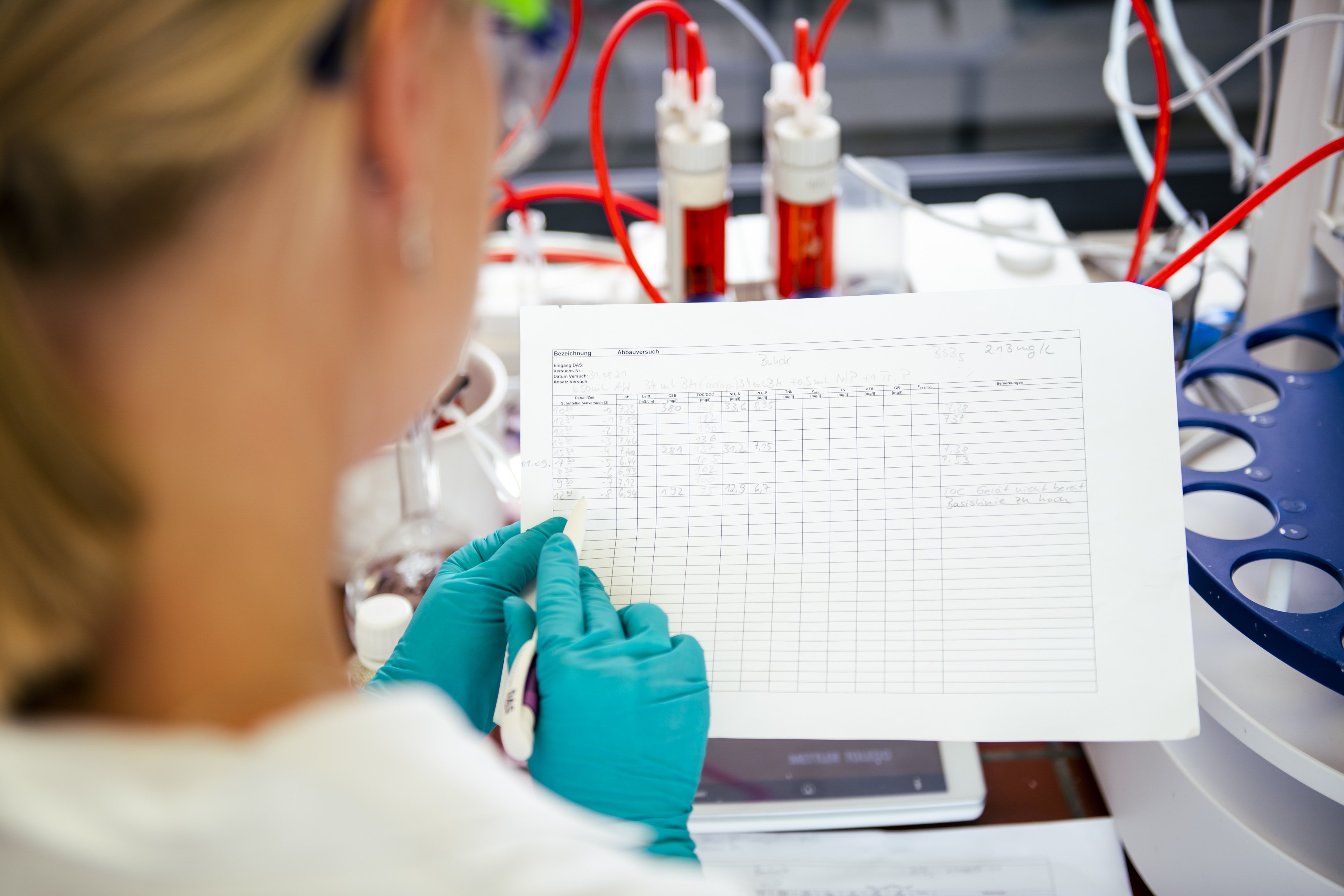 A scientist in a laboratory, wearing teal gloves, analyzes data on a sheet of paper while surrounded by scientific equipment and red tubing, reflecting a research environment.