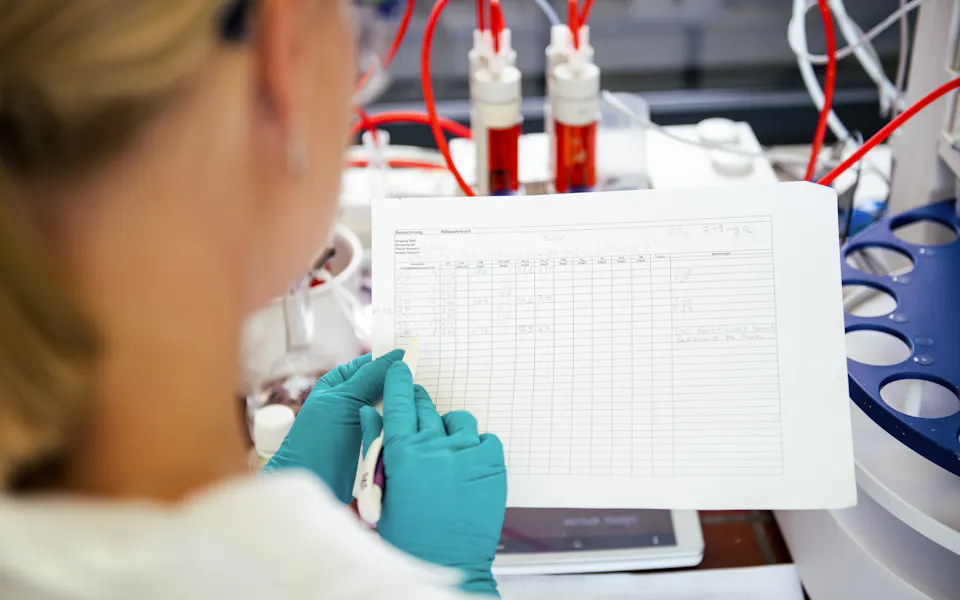 Documentation of laboratory tests A scientist in a laboratory, wearing teal gloves, analyzes data on a sheet of paper while surrounded by scientific equipment and red tubing, reflecting a research environment.