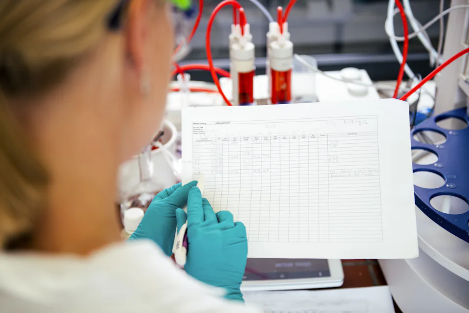 A scientist in a laboratory, wearing teal gloves, analyzes data on a sheet of paper while surrounded by scientific equipment and red tubing, reflecting a research environment.