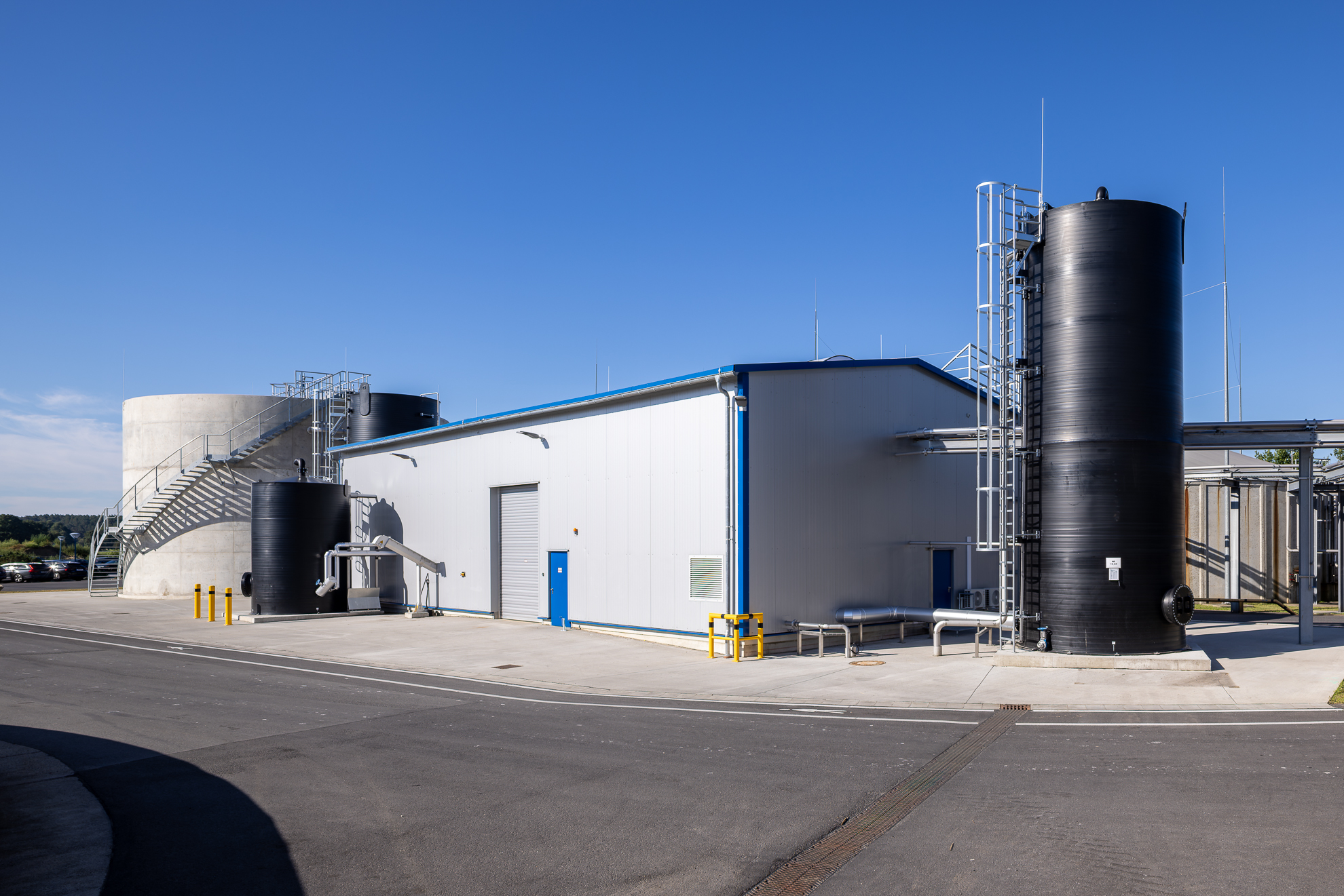 The image depicts a modern industrial facility with large cylindrical storage tanks and a white building, under a clear blue sky.
