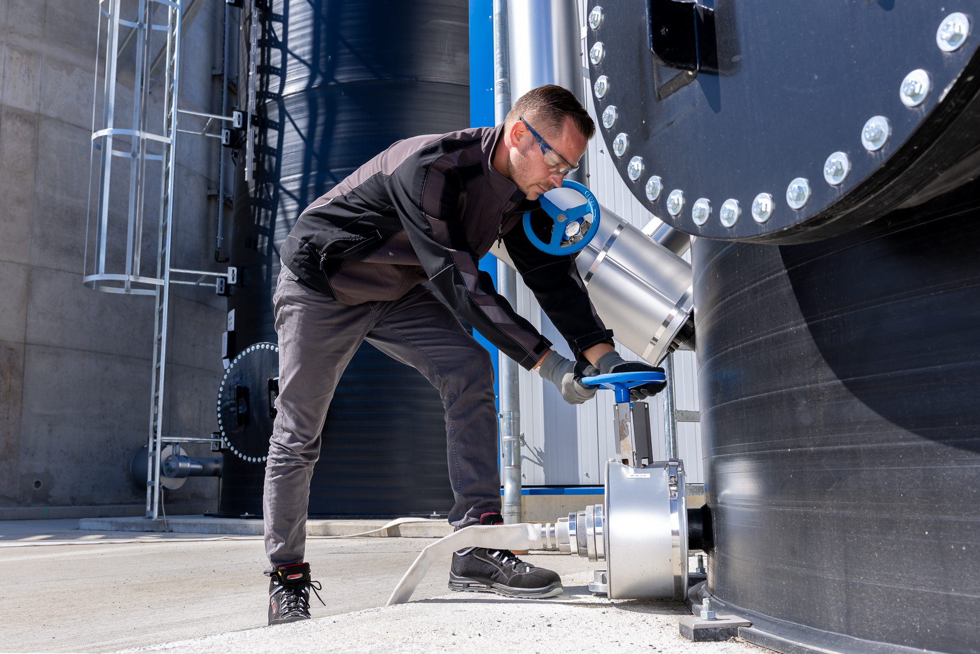 A person wearing safety glasses and gloves inspects a blue valve on an industrial metal storage tank, highlighting industrial safety and engineering processes.