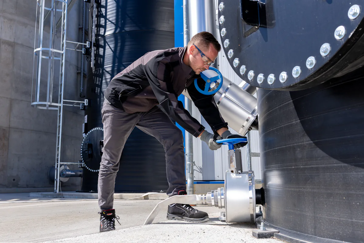 Emsland Aller Aqua MBBR Denitrification A person wearing safety glasses and gloves inspects a blue valve on an industrial metal storage tank, highlighting industrial safety and engineering processes.