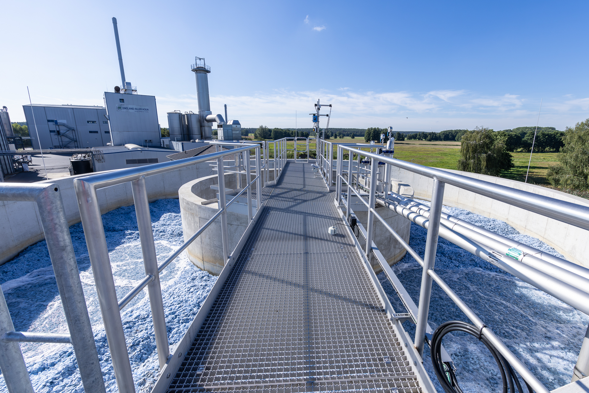 An aerial view of a modern water treatment plant featuring a steel walkway with railings, from which one can see circular basins filled with shimmering blue water, surrounded by industrial buildings and green fields on a clear, sunny day.