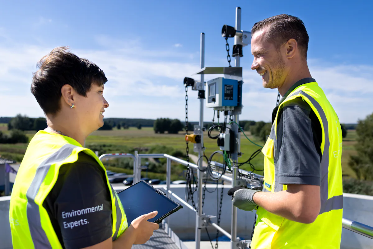 DAS Environmental Engineers Conducting Measurements Two people wearing high-visibility vests discuss atop an outdoor industrial platform with electronic equipment under a clear blue sky, embodying environmental monitoring and technological collaboration.