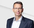 Portrait Thomas Schmidt, General Manager, AMTC Dresden A professionally dressed man wearing a navy suit and white shirt stands confidently against a minimalistic, light gray geometric background.