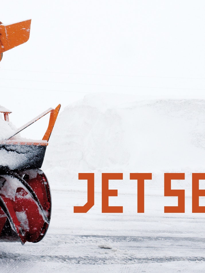 Snow blower with red auger ejecting snow, next to the text “JET SET” in orange letters on a white winter background.