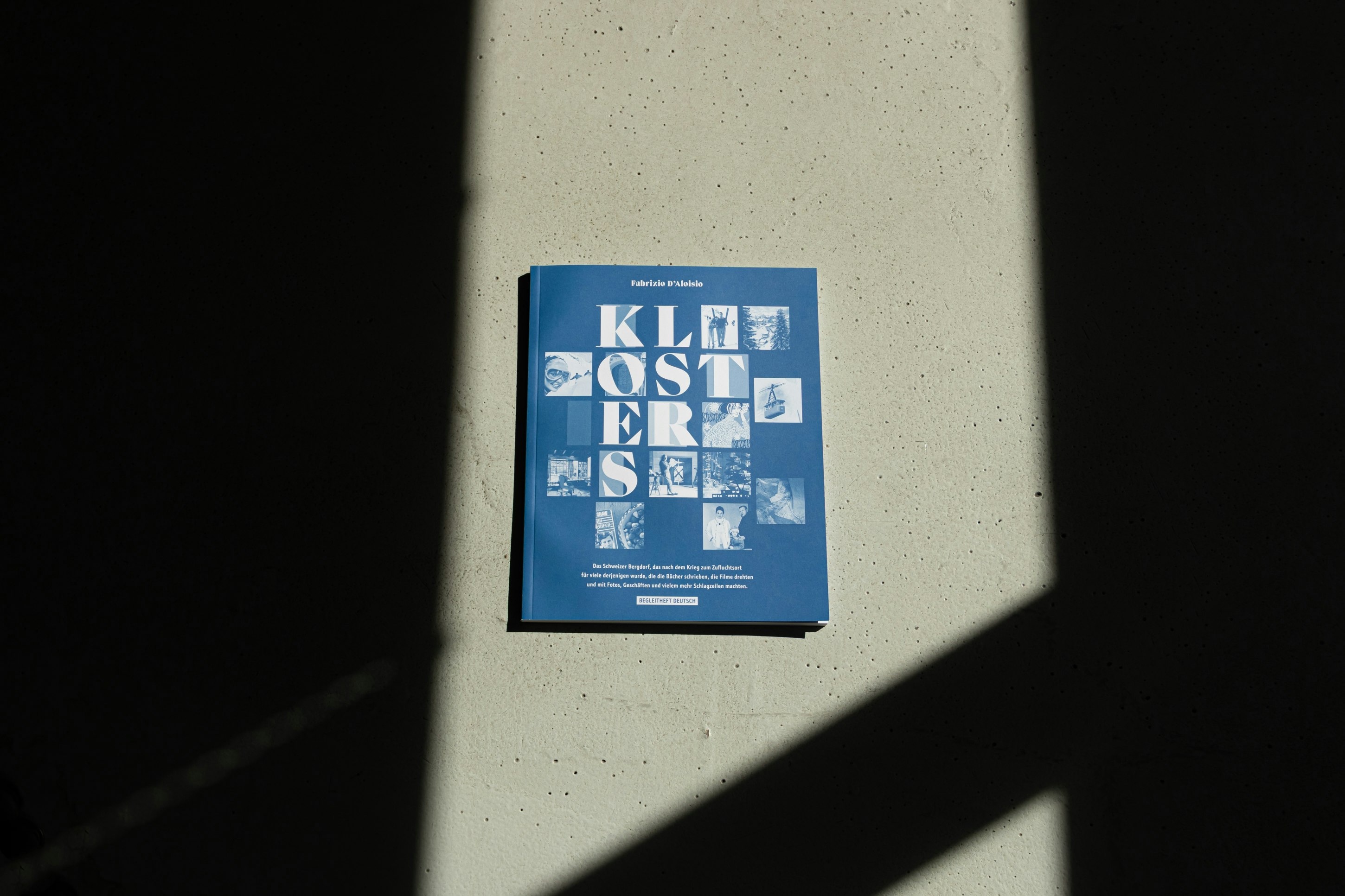 Blaues Buch „Klosters“ mit grafischem Cover, fotografiert im starken Licht- und Schattenspiel auf Betonboden.