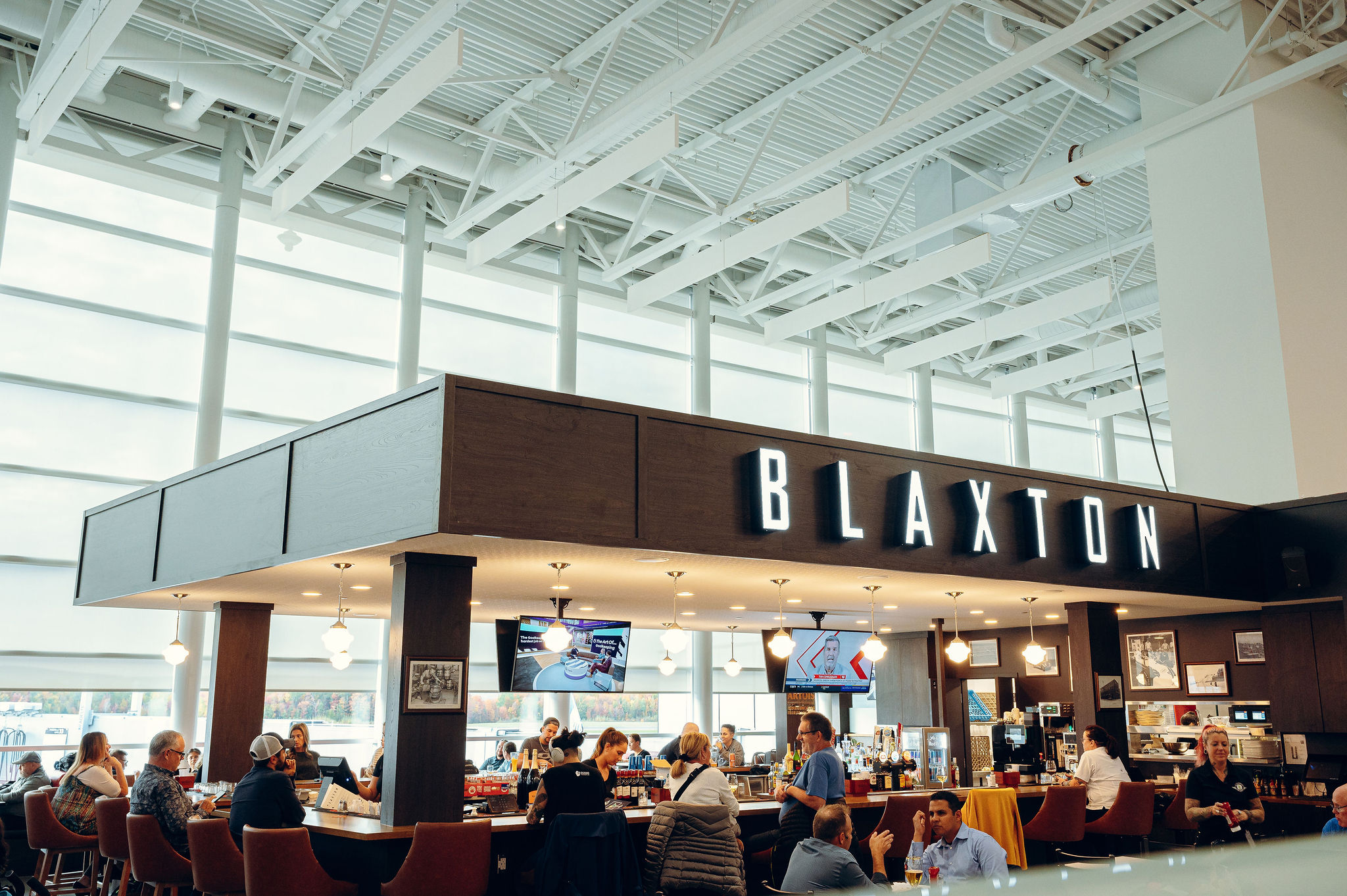 Les clients s'installent pour dîner dans un restaurant d'aéroport moderne appelé Blaxton, doté de grandes baies vitrées laissant entrer la lumière naturelle et de plusieurs téléviseurs visibles depuis la salle.