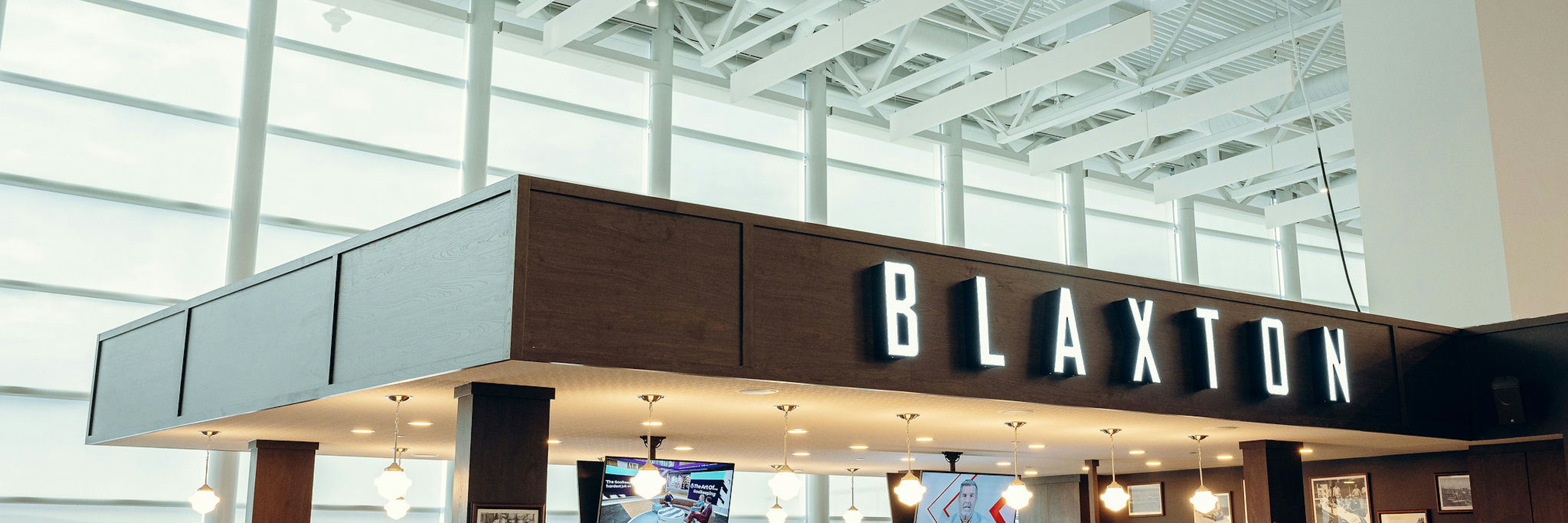 Les clients s'installent pour dîner dans un restaurant d'aéroport moderne appelé Blaxton, doté de grandes baies vitrées laissant entrer la lumière naturelle et de plusieurs téléviseurs visibles depuis la salle.