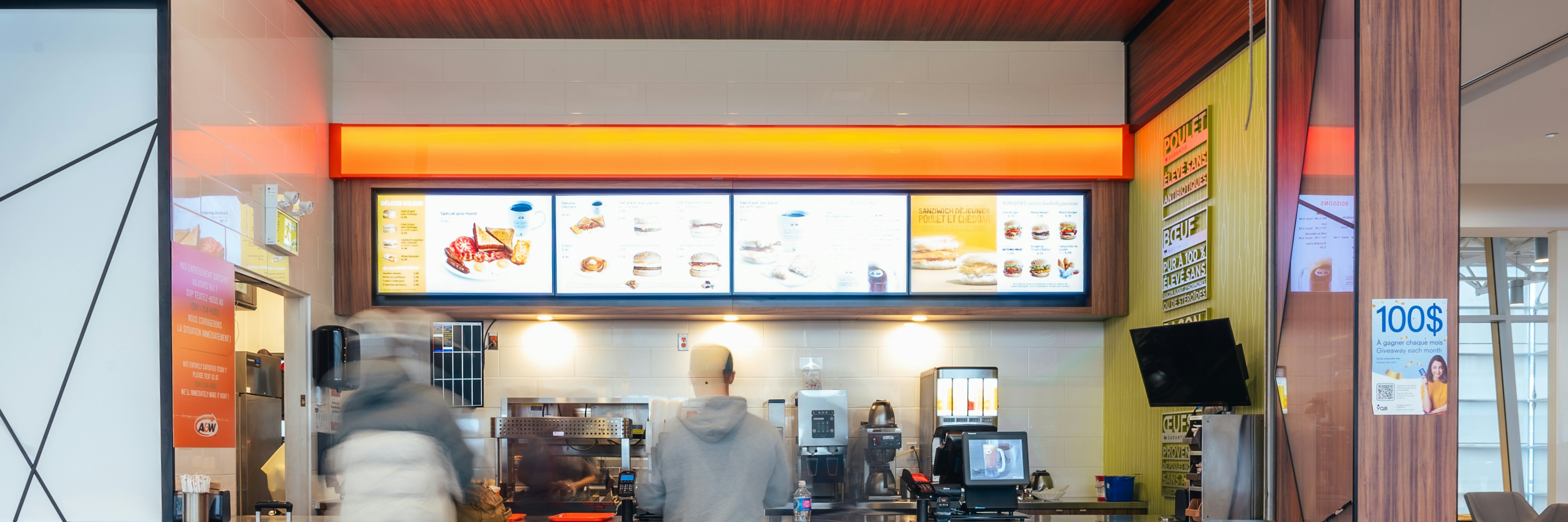 Two people stand at the counter of an A&W fast food restaurant, looking at the menu above. The area is roped off and brightly lit, with modern decor and digital menu screens.