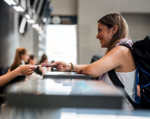 A woman carrying a backpack smiles as she hands her passport and ticket to a check-in agent at the airport, on the other side of the counter.