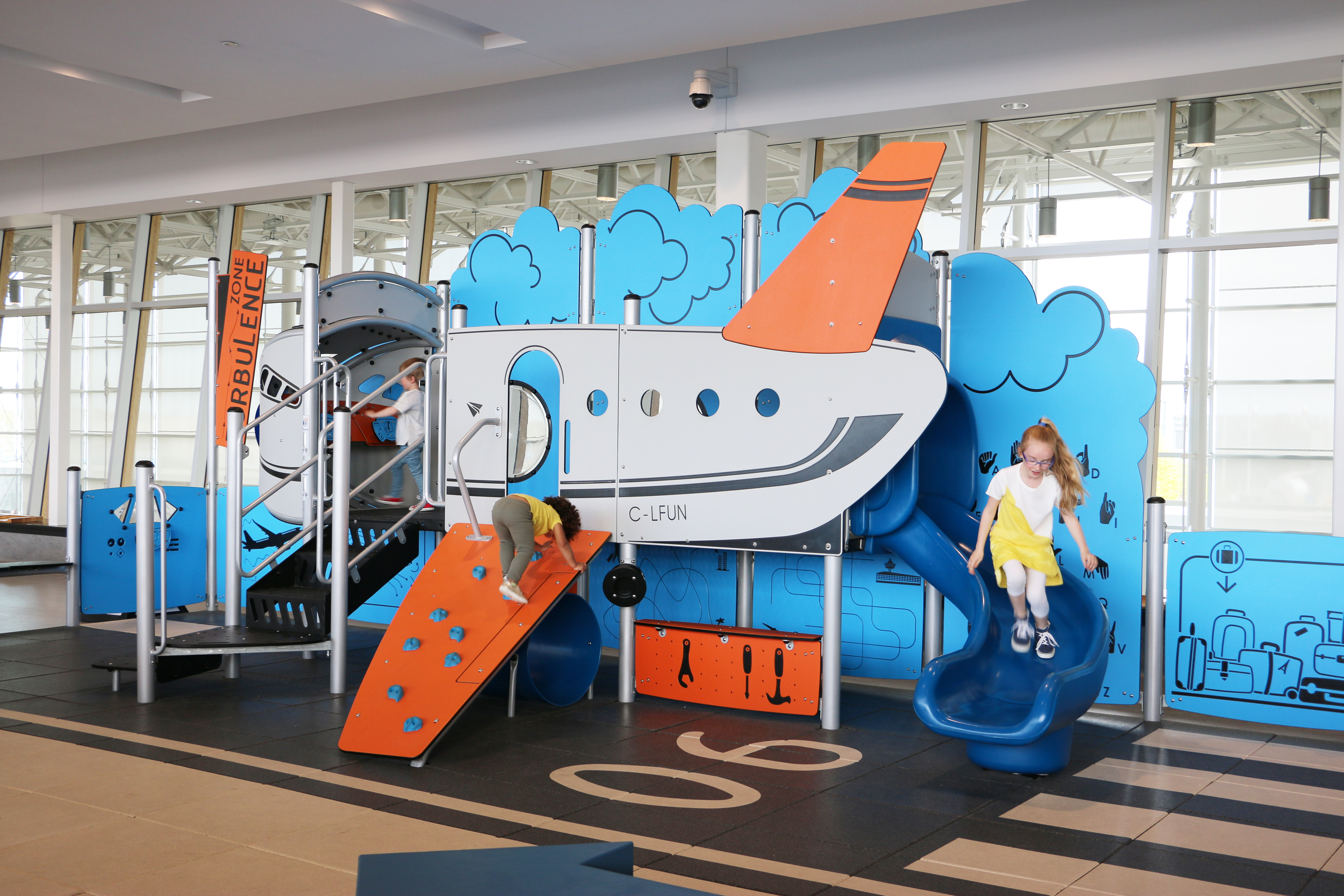 Deux enfants jouent dans une aire de jeux couverte sur le thème de l'avion, équipée d'un toboggan et d'un mur d'escalade. Des couleurs vives, bleu et orange, ornent la structure de jeux.